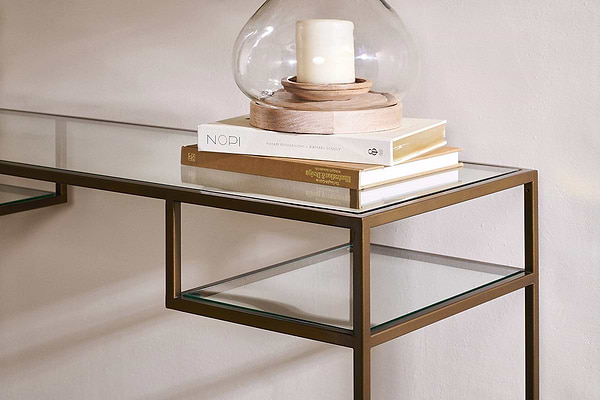 The nkuku Nakuru Iron & Glass Console Table features a bronze frame and glass top. Stacked books, including “NOPI,” sit atop it, alongside a round glass candle holder with a white pillar candle, set against a light-colored wall.