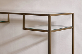 A close-up of the Nakuru Iron & Glass Console Table by nkuku shows its slim metallic bronze frame and clear glass shelves against a white wall, casting soft shadows. The minimalist design features clean, straight lines for a modern look.