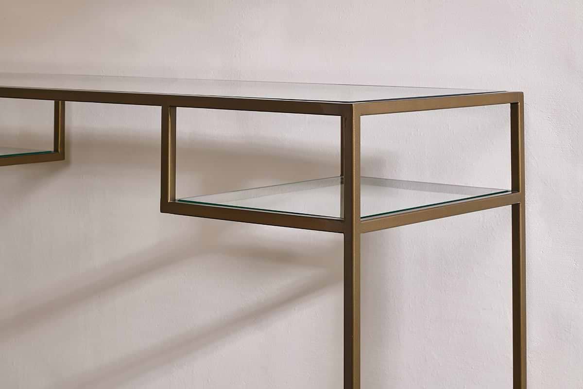 A close-up of the Nakuru Iron & Glass Console Table by nkuku shows its slim metallic bronze frame and clear glass shelves against a white wall, casting soft shadows. The minimalist design features clean, straight lines for a modern look.