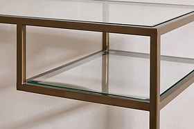 Close-up of the Nakuru Iron & Glass Console Table by nkuku, showcasing its bronze iron frame and clear glass surfaces. This contemporary hallway table features clean lines and a sleek look against a plain white wall.