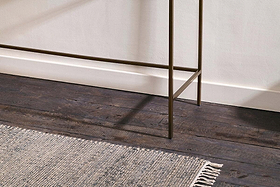 A close-up of dark wooden floorboards, a textured light gray fringed rug, white baseboards, and the slender bronze legs of the nkuku Nakuru Iron & Glass Console Table against a light-colored wall.