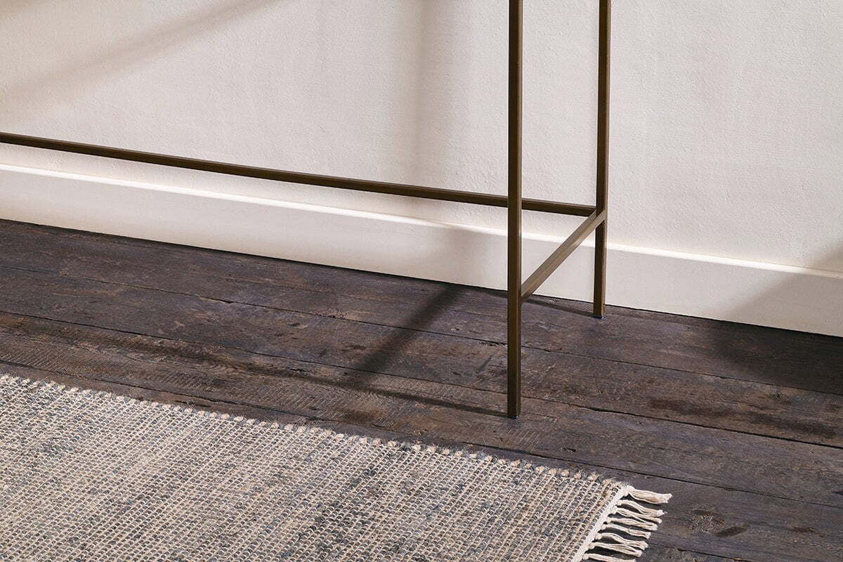 A close-up of dark wooden floorboards, a textured light gray fringed rug, white baseboards, and the slender bronze legs of the nkuku Nakuru Iron & Glass Console Table against a light-colored wall.