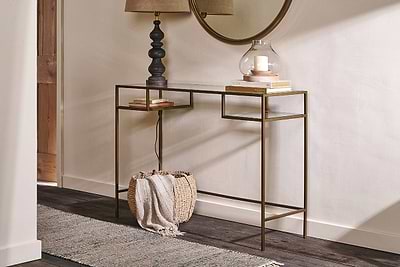 The Nakuru Iron & Glass Console Table by nkuku features a slim gold frame and glass top styled with a black lamp, glass candle holder, and books. A woven basket with a blanket sits below, while a round mirror and rug complete the look on dark wood floors.