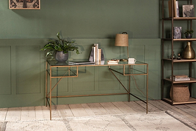 The Nakuru Iron & Glass Desk by nkuku, styled with a laptop, plant, lamp, and books, brings modern elegance to a home office set against a bold green paneled wall.