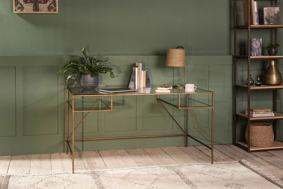 The Nakuru Iron & Glass Desk by nkuku, styled with a laptop, plant, lamp, and books, brings modern elegance to a home office set against a bold green paneled wall.