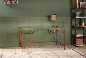 The nkuku Nakuru Iron & Glass Desk and a tan lamp stand out against a dark green paneled wall, creating an elegant home office setup. A tall shelving unit on the right displays decorative accents.