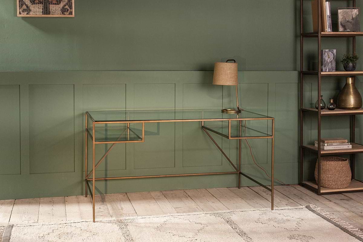 The nkuku Nakuru Iron & Glass Desk and a tan lamp stand out against a dark green paneled wall, creating an elegant home office setup. A tall shelving unit on the right displays decorative accents.