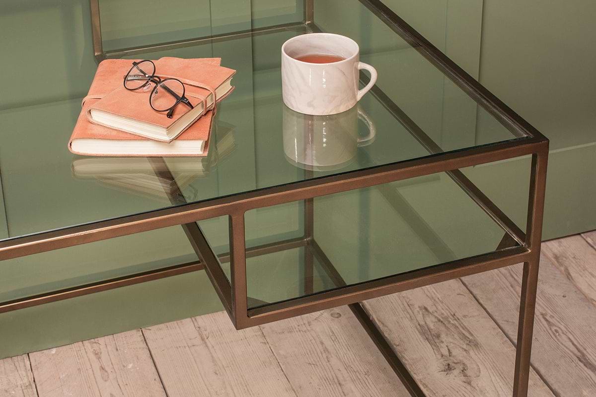 The Nakuru Iron & Glass Desk by nkuku features a closed notebook, glasses, and a white mug of tea on its glass top. The elegant desk stands on light wood flooring with a green wall in the background.