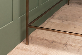 A close-up of the nkuku Nakuru Iron & Glass Desk showcases its bronze-finished metal leg on light wood flooring beside a green paneled wall. The visible wood grain and imperfections lend the scene a rustic charm.