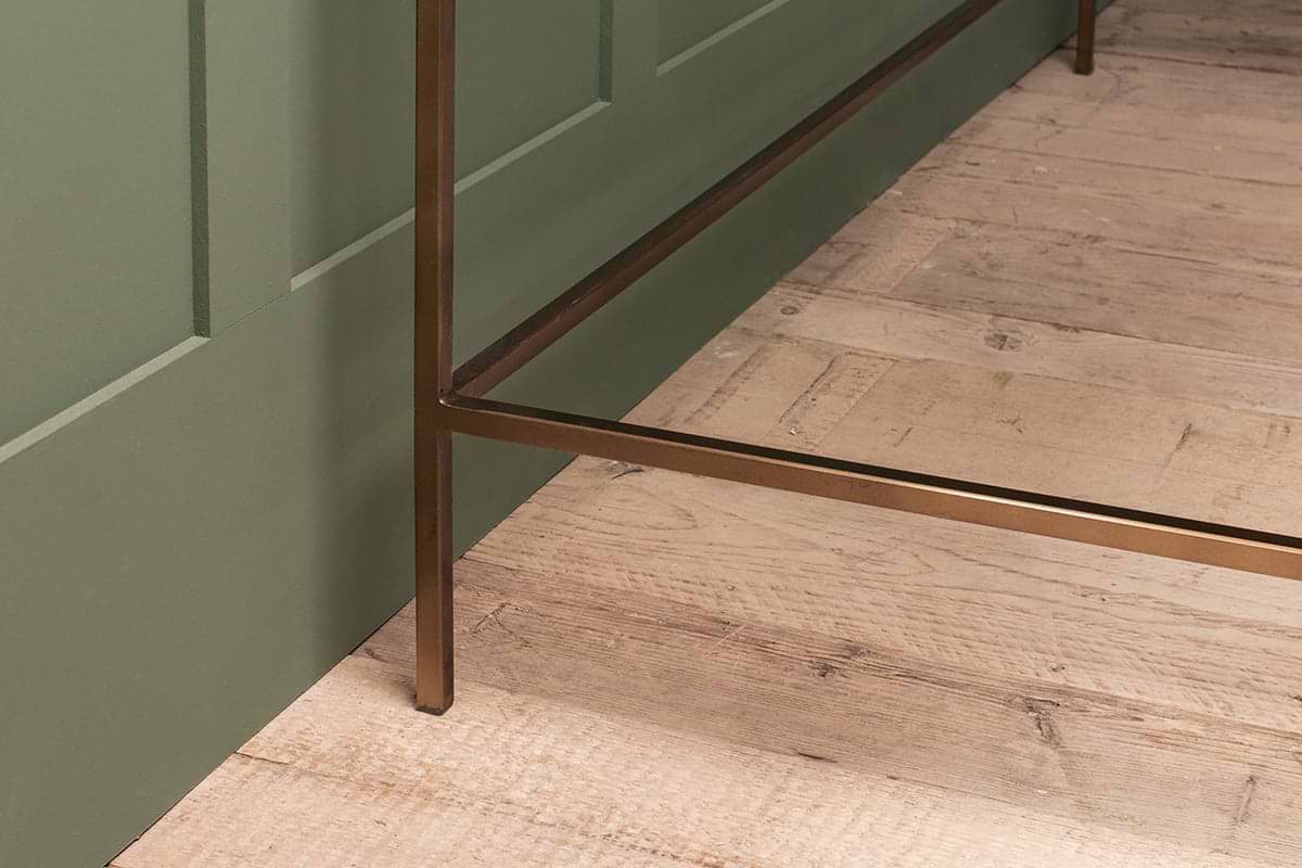 A close-up of the nkuku Nakuru Iron & Glass Desk showcases its bronze-finished metal leg on light wood flooring beside a green paneled wall. The visible wood grain and imperfections lend the scene a rustic charm.