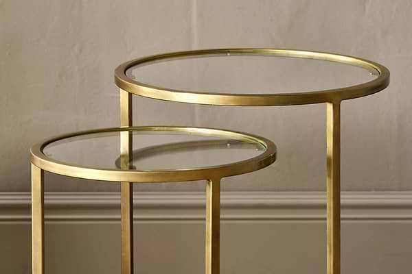 A pair of nkuku Nakuru Iron & Glass Side Table Set - Brass, featuring round glass tops and brass frames, sit elegantly against a textured wall and baseboard.
