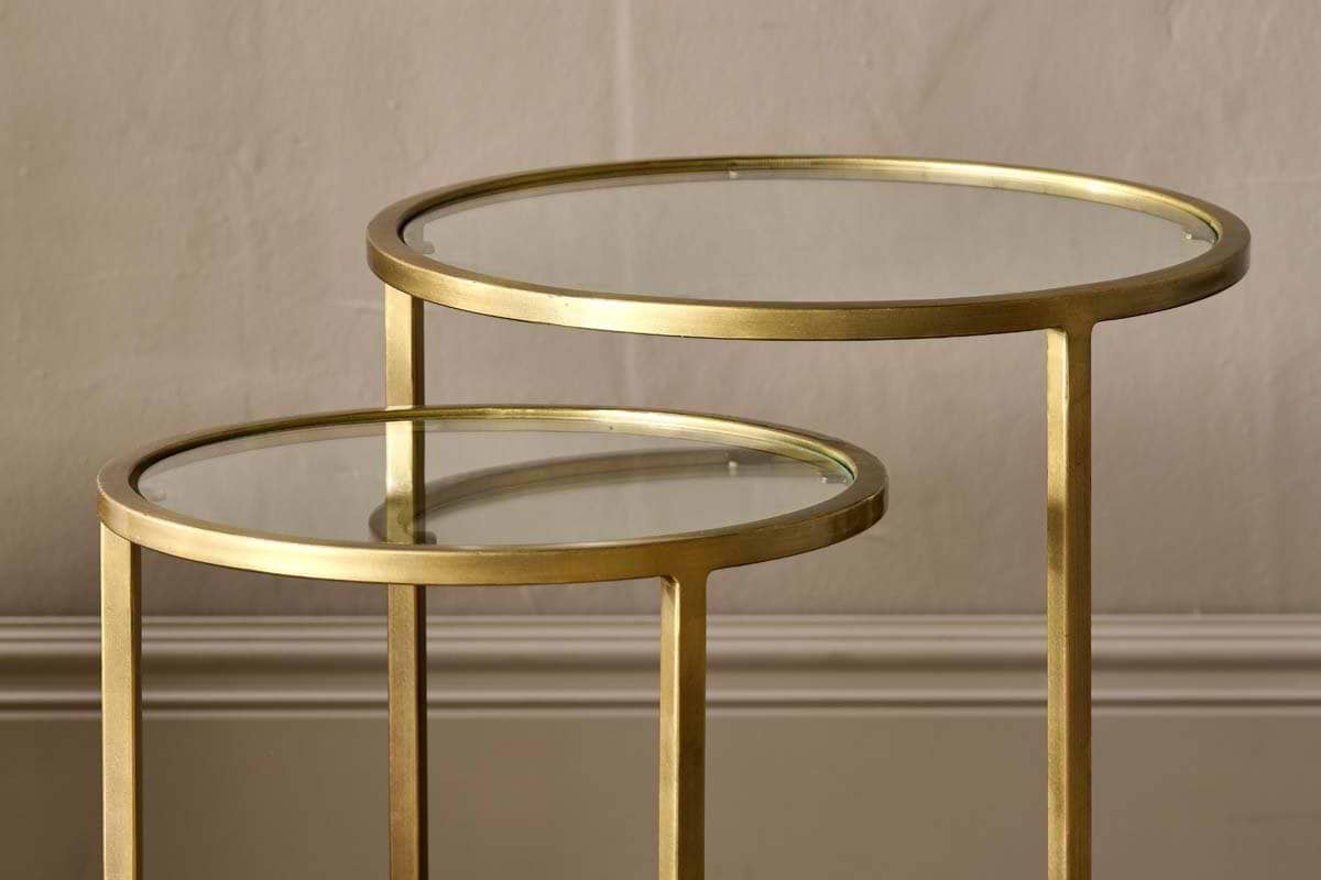 A pair of nkuku Nakuru Iron & Glass Side Table Set - Brass, featuring round glass tops and brass frames, sit elegantly against a textured wall and baseboard.