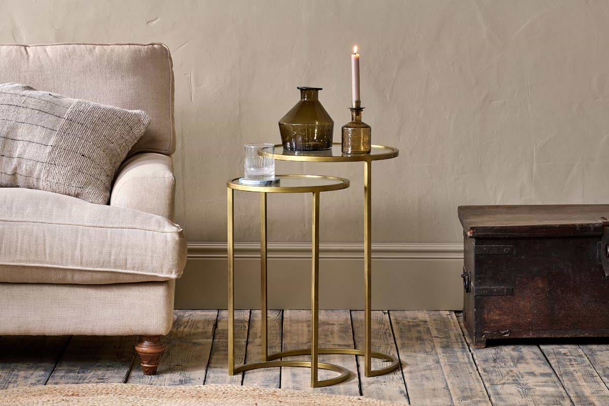 Two nested nkuku Nakuru Iron & Glass Side Tables in brass hold a glass of water, two brown vases, and a pink candle beside a beige sofa with a textured cushion, next to a dark wooden trunk on a rustic wooden floor.