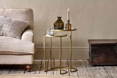 Two nested nkuku Nakuru Iron & Glass Side Tables in brass hold a glass of water, two brown vases, and a pink candle beside a beige sofa with a textured cushion, next to a dark wooden trunk on a rustic wooden floor.