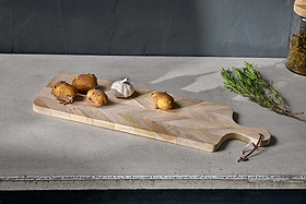 The nkuku Nalbari Chopping Board - Natural, crafted from sustainable mango wood with a chevron pattern, holds four unpeeled potatoes and a garlic bulb on a gray countertop beside rosemary and a jar of dried herbs.