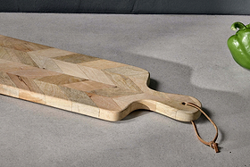 The nkuku Nalbari Chopping Board - Natural, crafted from sustainable mango wood with a chevron pattern and leather loop handle, rests on a light gray countertop. A green bell pepper is in the upper right; the background wall is dark gray.