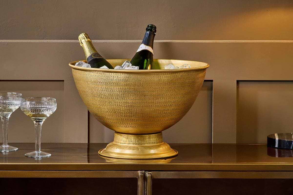 The nkuku Nalgora Wine & Champagne Bucket, with its large textured gold design, chills two bottles among ice. Two crystal coupe glasses rest nearby on a gleaming gold surface, evoking the spirit of a refined celebration.
