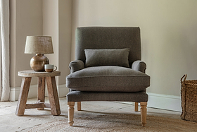 A cozy corner features the nkuku Nalla Wool Armchair in charcoal, accented by a herringbone cushion. The chair sits on a woven rug beside a rustic wooden side table with a ceramic lamp and cup, bathed in soft natural light through curtains.