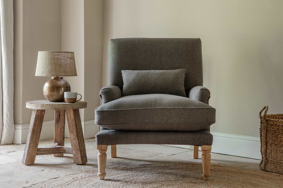 A cozy corner features the nkuku Nalla Wool Armchair in charcoal, accented by a herringbone cushion. The chair sits on a woven rug beside a rustic wooden side table with a ceramic lamp and cup, bathed in soft natural light through curtains.