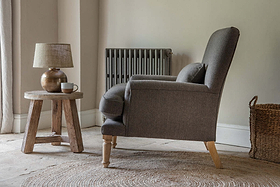 A cozy living room corner features the nkuku Nalla Wool Armchair in Charcoal, set on a round woven rug beside a rustic wooden side table topped with a ceramic lamp and cup. A gray radiator and woven basket complete the scene.