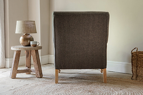The back of the nkuku Nalla Wool Armchair in charcoal is shown, with a small wooden stool holding a lamp and mug on its left, and a woven basket to the right of this cozy reading chair.