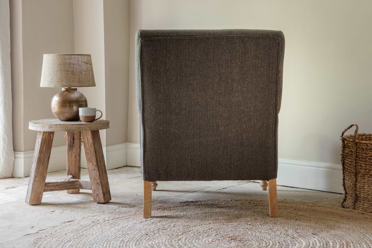 The back of the nkuku Nalla Wool Armchair in charcoal is shown, with a small wooden stool holding a lamp and mug on its left, and a woven basket to the right of this cozy reading chair.