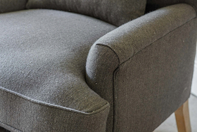 Close-up of the nkuku Nalla Wool Armchair in Charcoal, featuring a subtle herringbone pattern, curved armrest, seat cushion, and wooden leg. The soft wool fabric and modern design make it an inviting choice for reading.