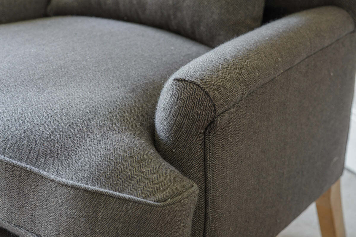 Close-up of the nkuku Nalla Wool Armchair in Charcoal, featuring a subtle herringbone pattern, curved armrest, seat cushion, and wooden leg. The soft wool fabric and modern design make it an inviting choice for reading.