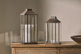 Two nkuku Nampo Lanterns in Aged Antique brass sit on a wooden sideboard—a tall lantern with a large white pillar candle, and a shorter one holding a tealight—casting soft shadows on the beige wall behind them.