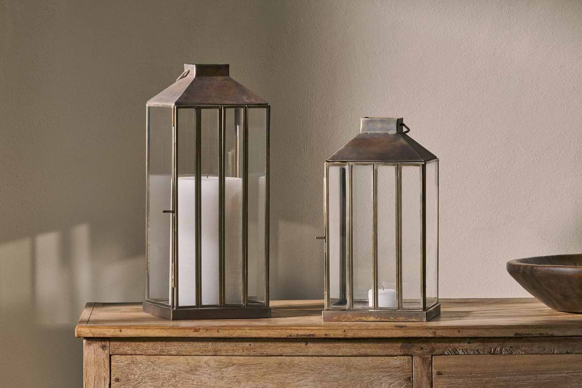 Two nkuku Nampo Lanterns in Aged Antique brass sit on a wooden sideboard—a tall lantern with a large white pillar candle, and a shorter one holding a tealight—casting soft shadows on the beige wall behind them.