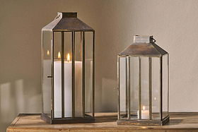 Two nkuku Nampo Lanterns - Aged Antique, one tall with a pillar candle and one short with a tealight, cast warm light from their brass and glass frames onto a wooden surface against a beige wall.