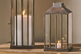 Two nkuku Nampo Lanterns - Aged Antique, made of brass and glass, sit on wood. The left contains a large white pillar candle with a tall flame; the right holds a smaller lit candle, both casting a warm glow against a neutral background.