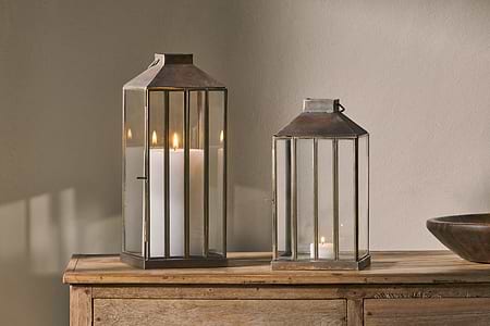 Two nkuku Nampo Lanterns - Aged Antique, one tall with a pillar candle and one short with a tealight, sit on a wooden surface. Warm candlelight glows through the brass and glass, casting soft shadows on the wall behind them.