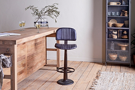 A modern kitchen featuring an nkuku Narwana Bar Chair in aged black, a wooden table by a light wall, two glasses of white wine, a vase with greenery, a glass cabinet with dishes, and a beige textured rug on wooden flooring.