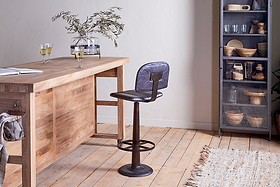A rustic wooden island bar features the nkuku Narwana Bar Chair in aged black. Champagne glasses and a vase with branches sit atop the bar, while a metal cabinet filled with kitchenware stands to the right.
