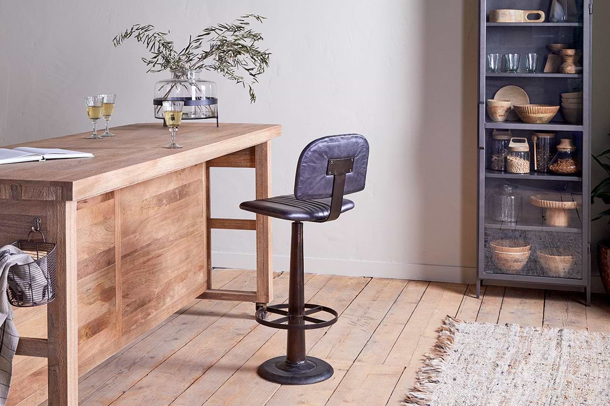 A rustic wooden island bar features the nkuku Narwana Bar Chair in aged black. Champagne glasses and a vase with branches sit atop the bar, while a metal cabinet filled with kitchenware stands to the right.