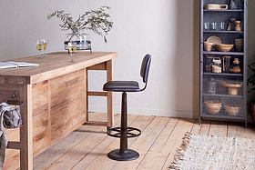 A modern wooden table with a glass vase and two white wine glasses sits beside the nkuku Narwana Bar Chair in aged black. A cabinet with glass doors holds dishes and baskets, while light wood floors and a beige rug finish the room.