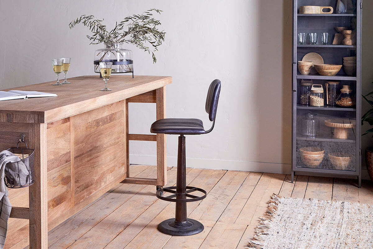 A modern wooden table with a glass vase and two white wine glasses sits beside the nkuku Narwana Bar Chair in aged black. A cabinet with glass doors holds dishes and baskets, while light wood floors and a beige rug finish the room.