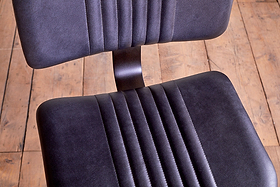 Close-up of the nkuku Narwana Bar Chair - Aged Black, a retro-style leather seat with vertical stitching on seat and backrest, shown on a light wooden floor with visible grain and gaps between planks.