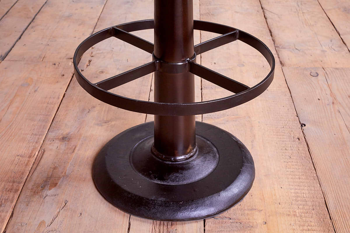 A close-up of the Narwana Bar Chair - Aged Black by nkuku shows its metal base with a black circular footrest and central support, standing on worn, light brown wooden planks with visible grain and knots.