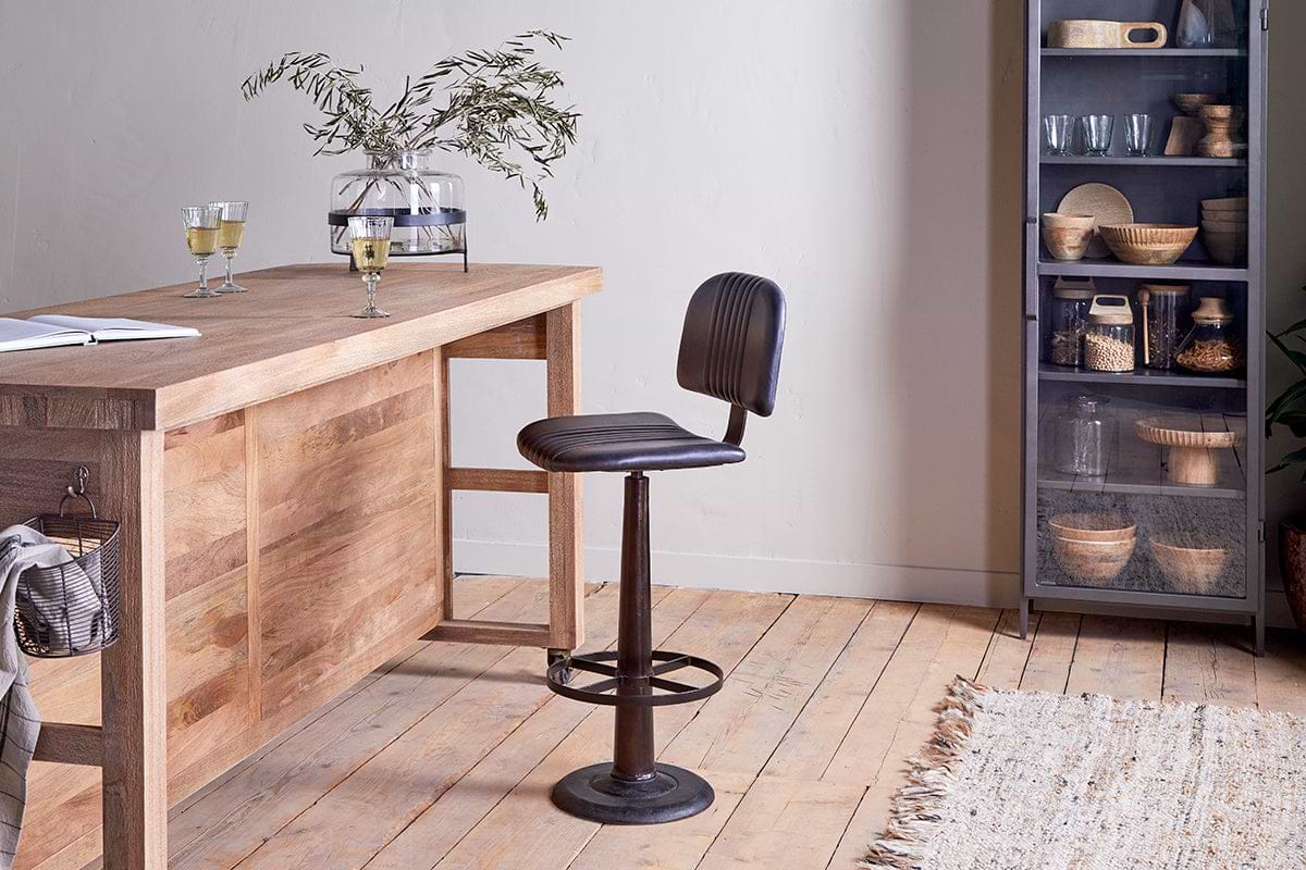 A modern kitchen features a wooden table with two glasses of white wine and a vase of greenery. Nearby stands the nkuku Narwana Bar Chair in Aged Black. Shelves hold dishes and jars, while a woven rug decorates the wooden floor.