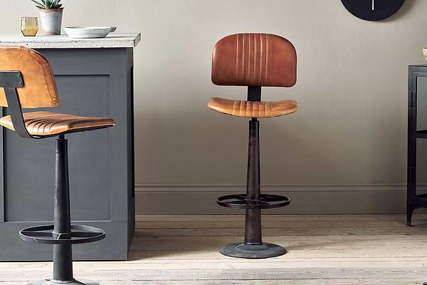 The Narwana Bar Chair - Aged Tan by nkuku, featuring brown leather and a black metal base with footrest, stands on a light wooden floor near a grey kitchen island. Another similar chair is partly visible on the left side of the image.