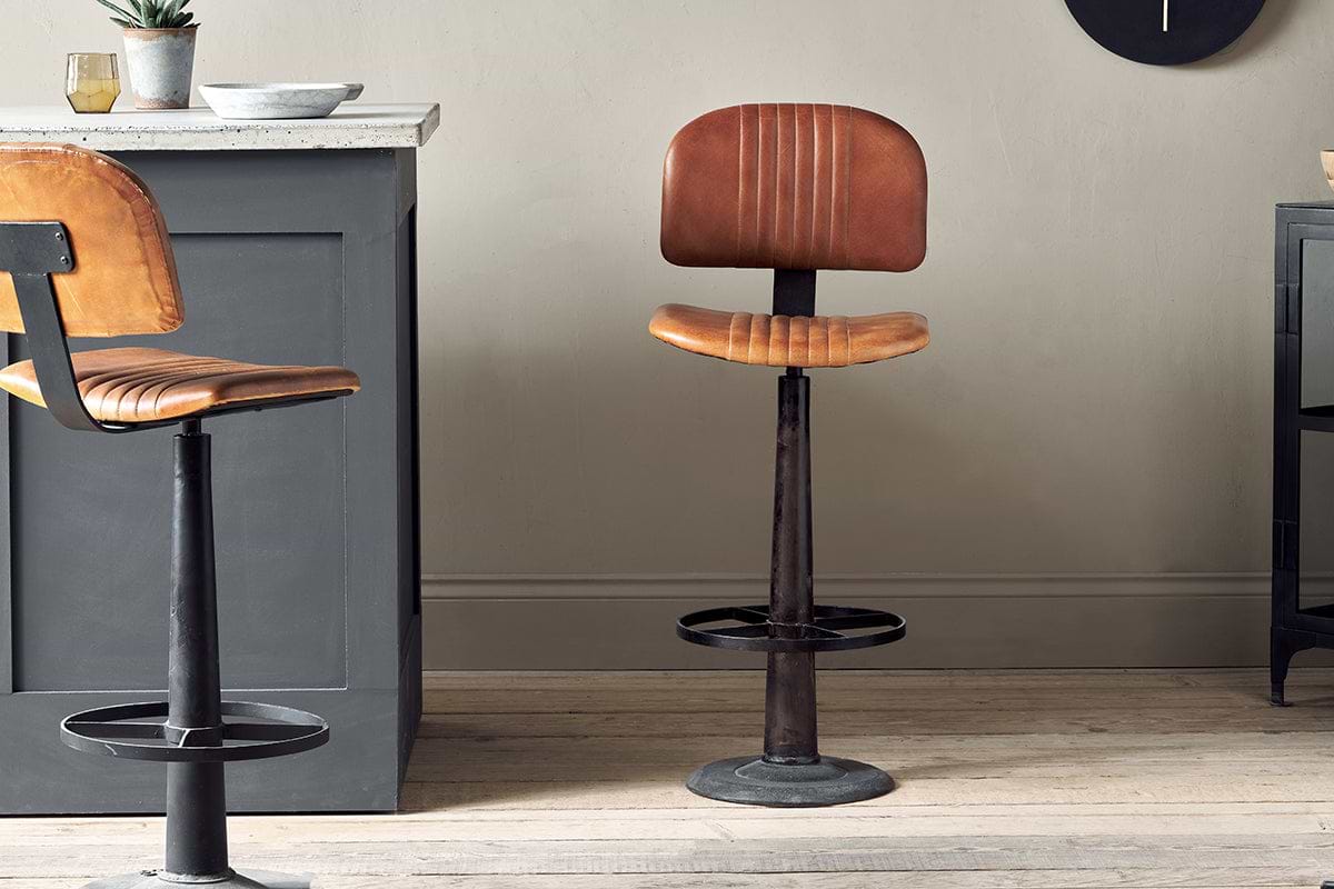 The Narwana Bar Chair - Aged Tan by nkuku, featuring brown leather and a black metal base with footrest, stands on a light wooden floor near a grey kitchen island. Another similar chair is partly visible on the left side of the image.
