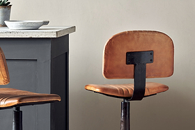 A modern kitchen with a gray island and concrete countertop features two nkuku Narwana Bar Chairs in aged tan leather with visible seams, metal frames, and cushioned seats and backs. A bowl rests on the countertop.