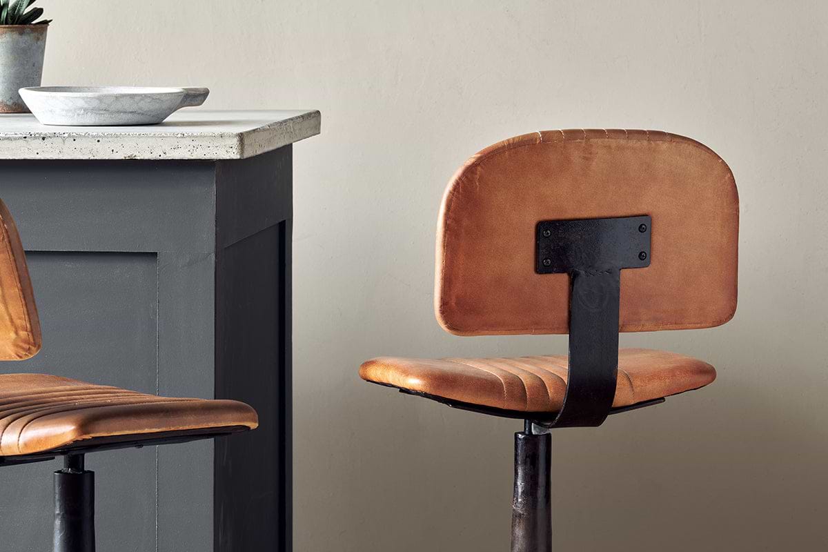 A modern kitchen with a gray island and concrete countertop features two nkuku Narwana Bar Chairs in aged tan leather with visible seams, metal frames, and cushioned seats and backs. A bowl rests on the countertop.
