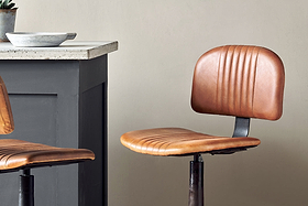 The Narwana Bar Chair - Aged Tan by nkuku, featuring a ribbed leather seat and backrest, sits beside a dark gray kitchen counter with a light stone top and matching metal base. A bowl is placed on the countertop.