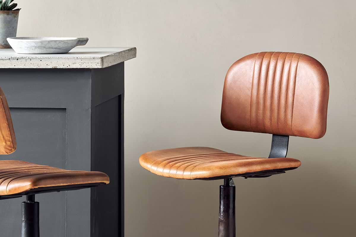 The Narwana Bar Chair - Aged Tan by nkuku, featuring a ribbed leather seat and backrest, sits beside a dark gray kitchen counter with a light stone top and matching metal base. A bowl is placed on the countertop.