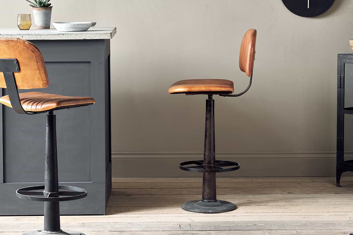 The Narwana Bar Chair - Aged Tan by nkuku, featuring brown leather and a metal base with a circular footrest, sits by a gray kitchen island. Another similar stool is partially visible on the left. The room has light wood floors and beige walls.