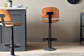 The Narwana Bar Chair - Aged Tan by nkuku, featuring brown leather and a black metal base, stands on a light wood floor beside a gray kitchen island in a modern, minimalistic setting. A matching stool is partially visible on the left.