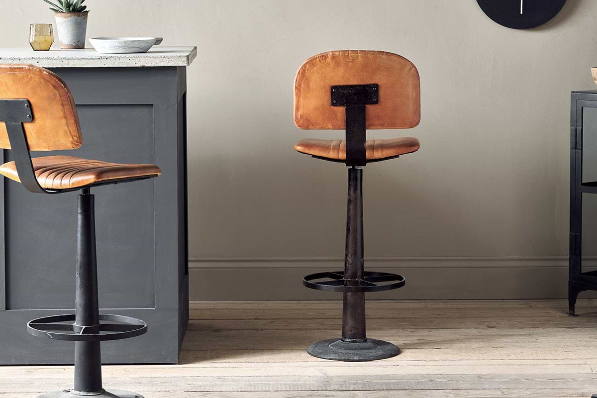 The Narwana Bar Chair - Aged Tan by nkuku, featuring brown leather and a black metal base, stands on a light wood floor beside a gray kitchen island in a modern, minimalistic setting. A matching stool is partially visible on the left.